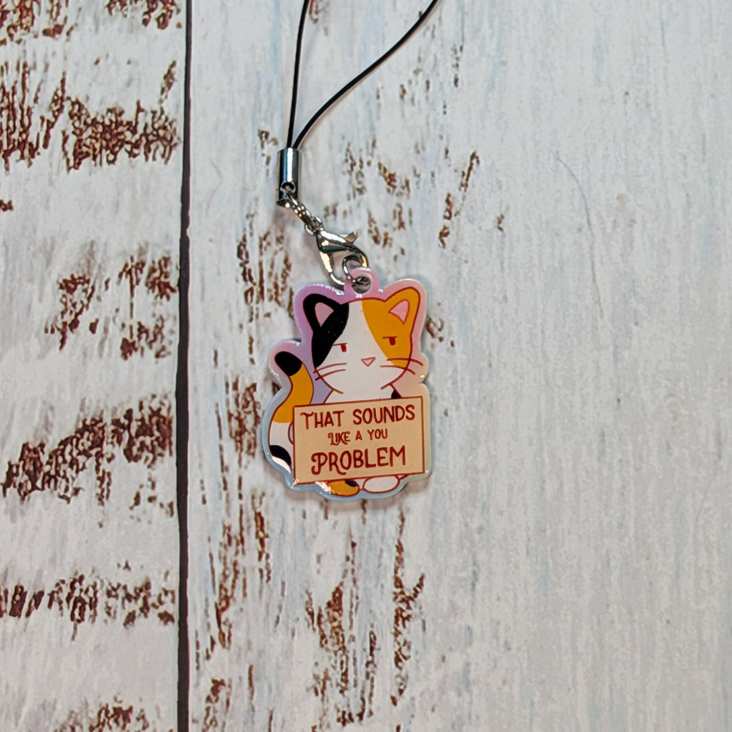 A small acrylic charm keychain featuring a cartoon cat with the text 'THAT SOUNDS WAY TOO PROBLEM', hanging from a black cord on a distressed white wooden surface.