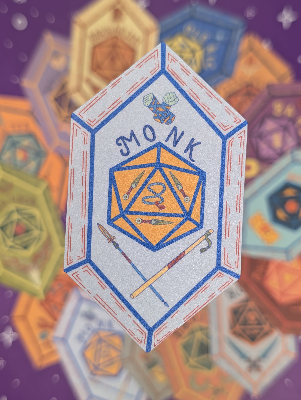 A hexagonal illustrated sticker featuring a white background, blue border, and an orange-yellow 20-sided die with a snake and the word "MONK" above it. The patch is centered against a blurred purple background filled with other colorful geometric icons.
