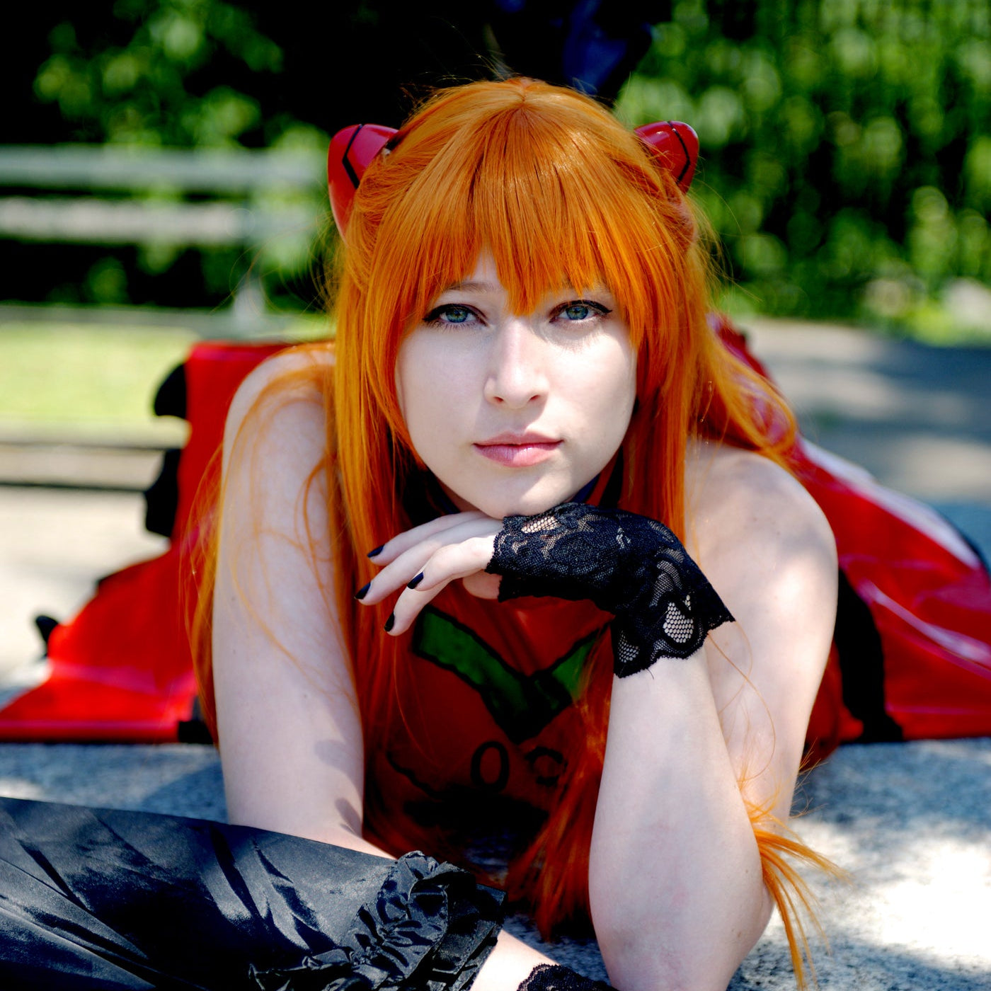 A woman in a detailed anime cosplay costume, featuring bright orange hair, a red top with a green emblem, and black lace fingerless gloves, lying on a stone surface outdoors.