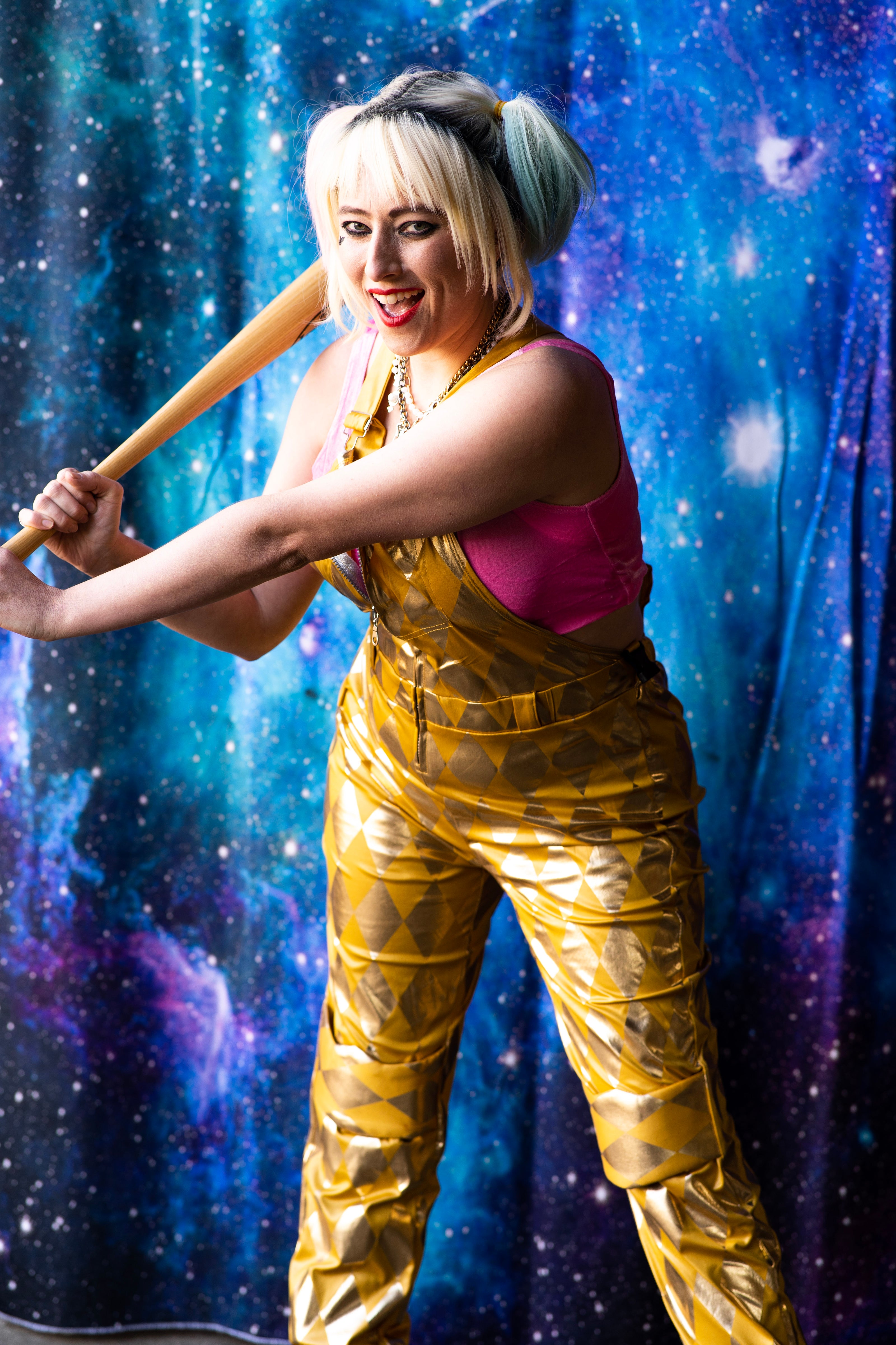 A woman in a metallic gold Harley Quinn costume with a pink top, holding a wooden baseball bat against a blue and purple starry space backdrop.