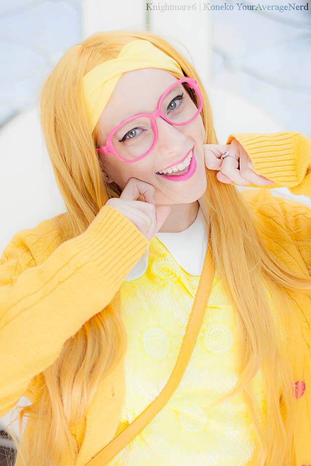 A woman wearing a yellow costume with a matching yellow headband, pink glasses, and a yellow cardigan over a white collared shirt, posing in a bright indoor setting.