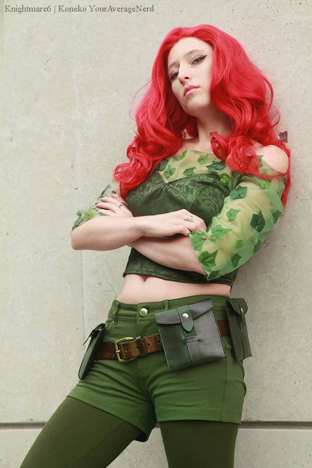 A woman with long bright red hair poses in a Poison Ivy cosplay outfit, featuring a green and yellow leaf-patterned crop top, green shorts, and a matching utility belt, set against a neutral concrete wall.