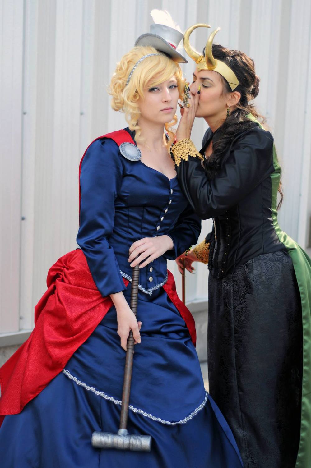 Two women in elaborate costumes posing outdoors. The woman on the left wears a blue and red Thor-inspired dress with a hammer prop and a hat, while the woman on the right wears a black and green Loki-inspired costume with a gold horned headpiece and leans in to whisper.