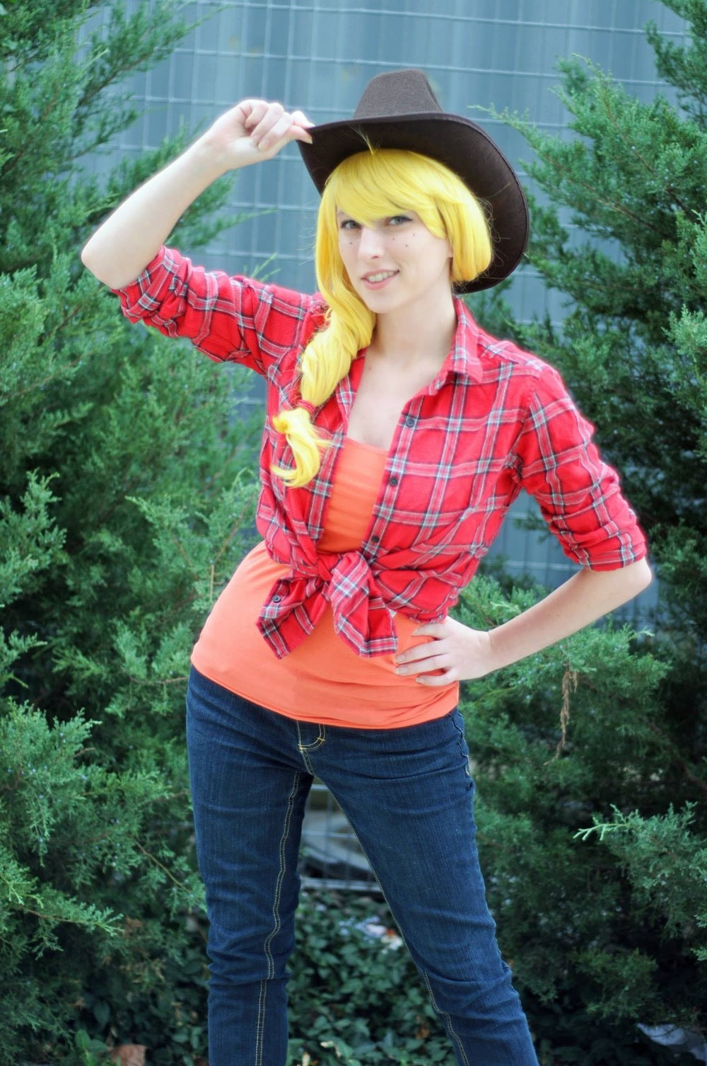 A woman posing outdoors in a costume consisting of a red and black plaid shirt tied at the waist, an orange top underneath, blue jeans, a brown cowboy hat, and a bright yellow wig.