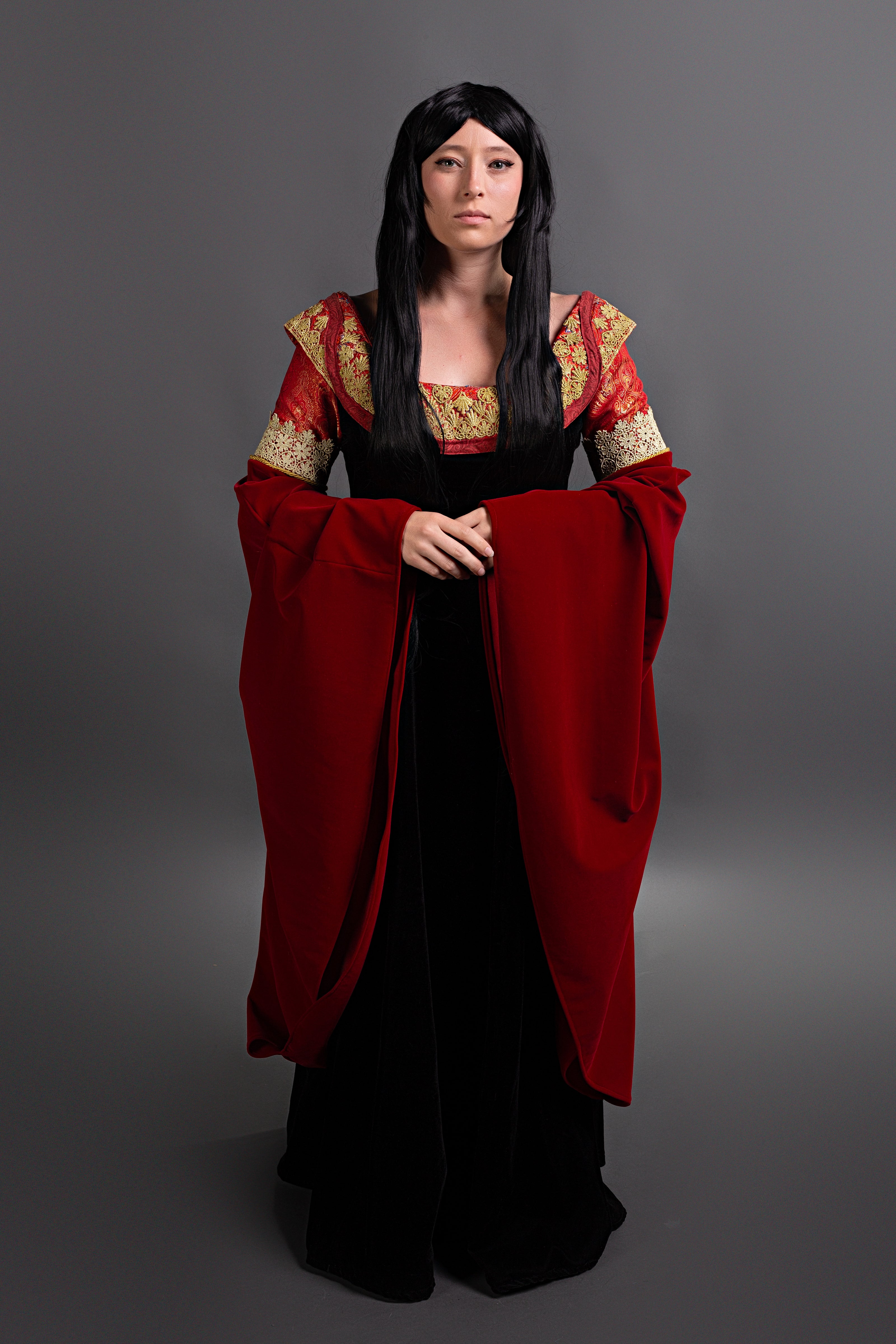 A woman modeling a medieval-style costume consisting of a black dress with a red velvet sleeves and a red and gold patterned bodice, set against a dark gray studio background.