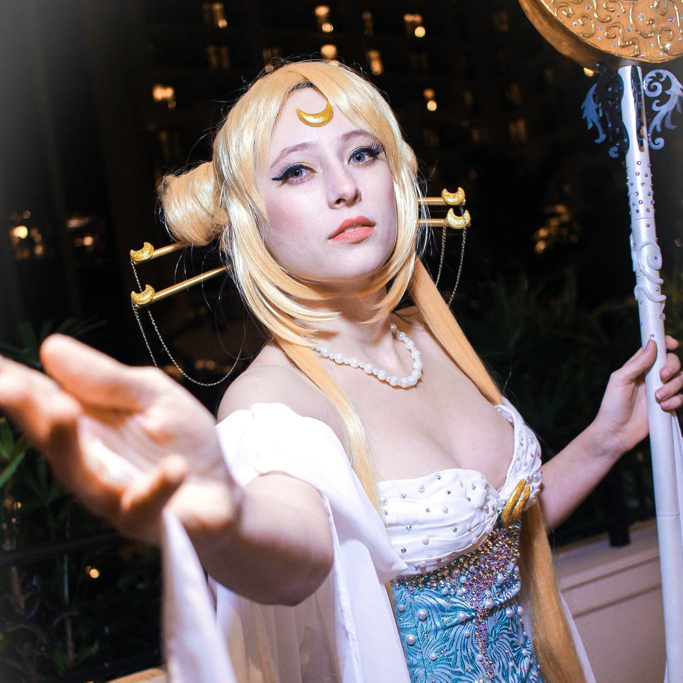 A woman in a detailed cosplay costume posing at night. The costume features a white and light blue patterned dress with a pearl necklace, a blonde wig with twin buns and a crescent moon headpiece, and a large golden staff held in one hand.