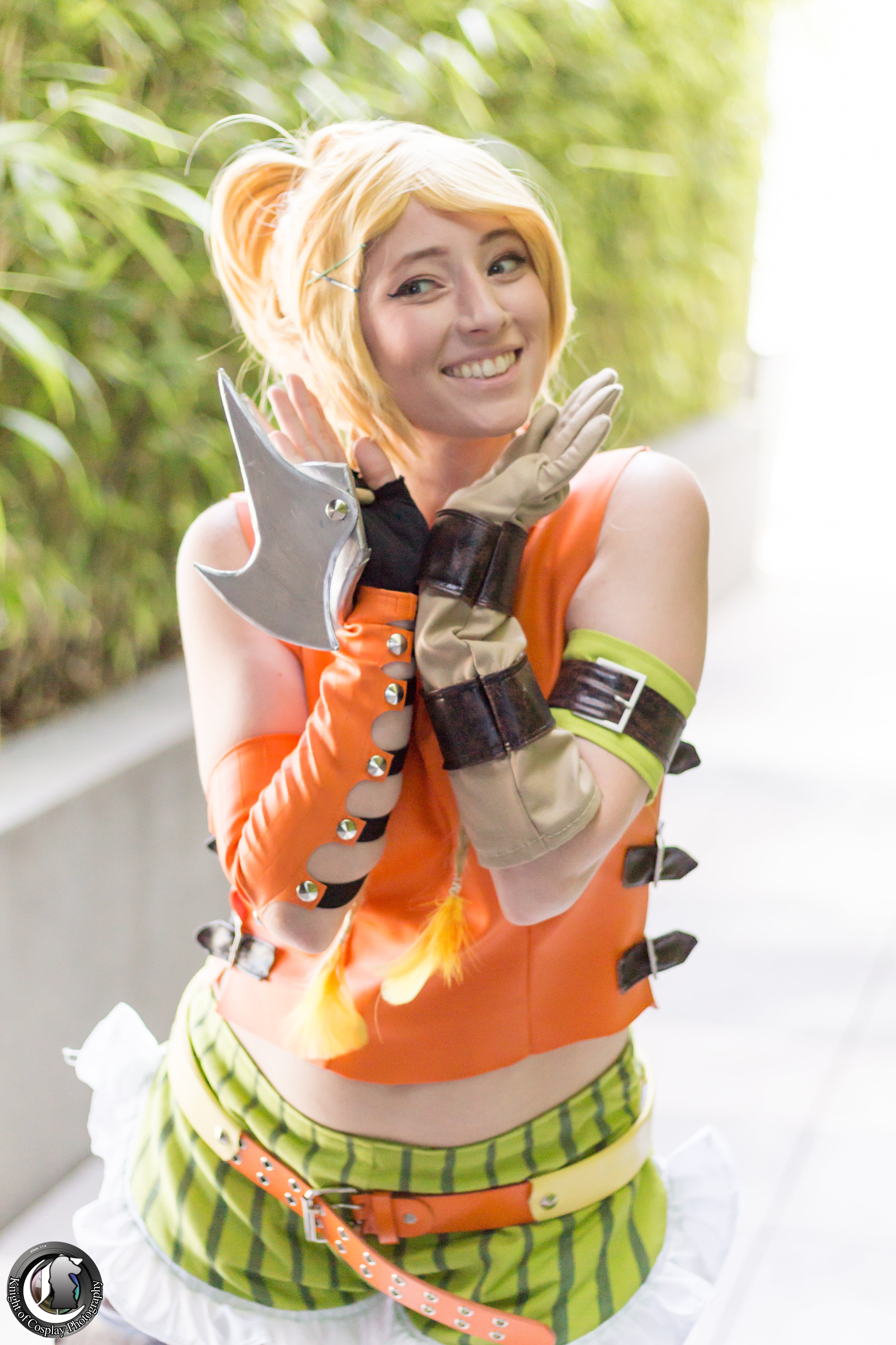 A woman posing in a detailed anime cosplay outfit, featuring a bright orange top, green and white striped skirt, and holding a silver double-bladed axe prop.