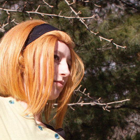 A person outdoors wearing a short strawberry blonde wig with a black headband and a pale yellow top, set against a background of pine trees and bare branches.